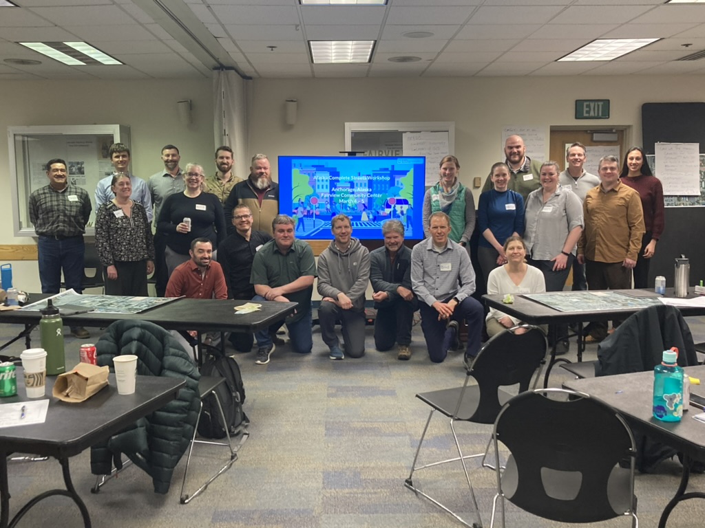 A group photo during a workshop in Alaska. Two rows of people are divided by a TV screen that displays a presentation. 