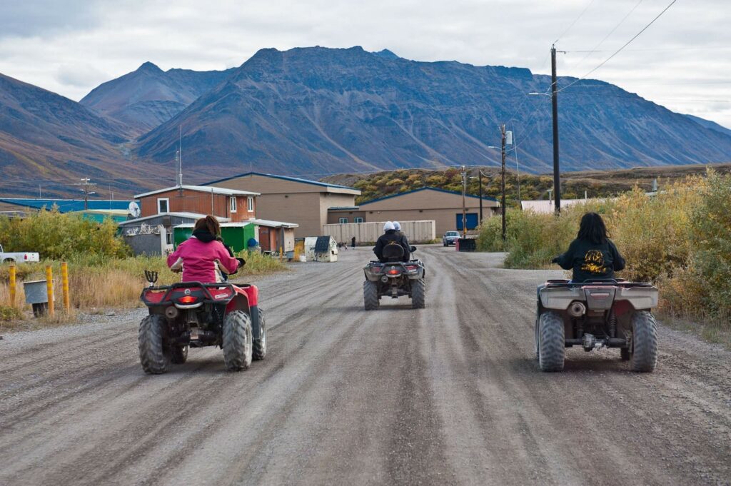 Three people ride on ATV's on a dirt road with mountains in the background.