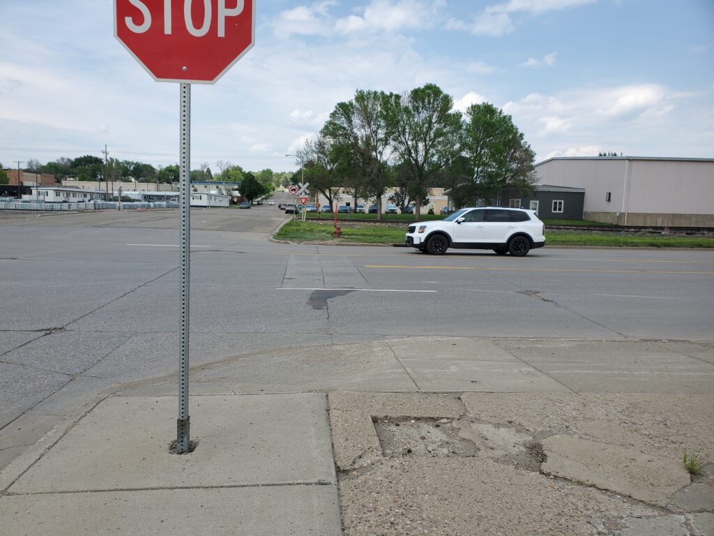 Photograph of a wide road with no crosswalk, and only a stop sign to signal to drivers to stop and wait for pedestrians to cross.