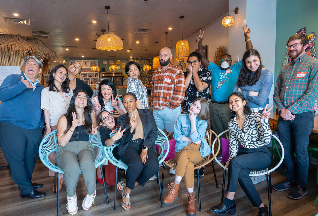 A diverse group of smiling individuals, seated and standing, making silly expressions for a group photo inside a restaurant.