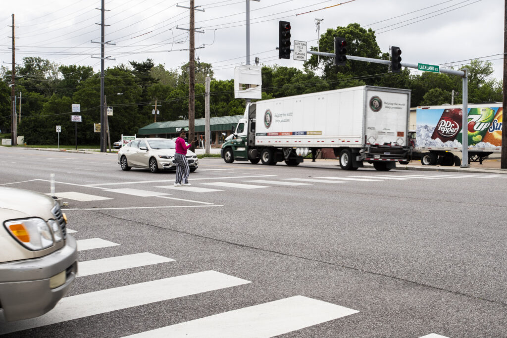 A photo taken on the corner of Lackland and 16th St. intersection in St. Louis, MO, where a pedestrian can be seen using a crosswalk as a semi truck drives by. 