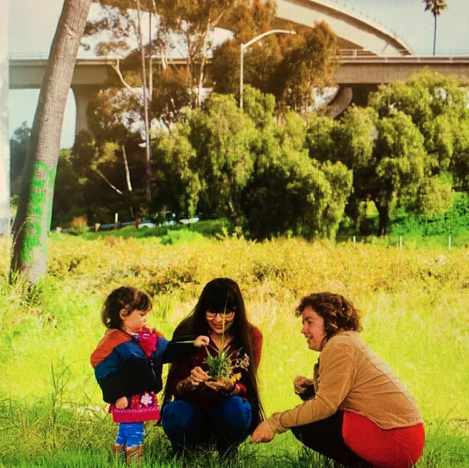 Two women crouch in front of a child playing in the grass, with a raised highway looming in the background