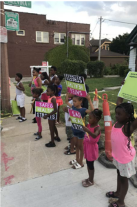 Ten young children in pink and white shirts hold up pink and green signs calling for safe, healthy streets.