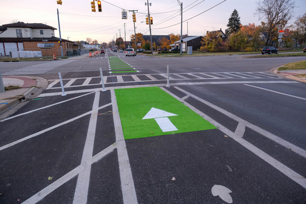 A painted bike lane and flex pots help reclaim space for cyclists while a marked crosswalk and traffic light signal for cars to stop for pedestrians.