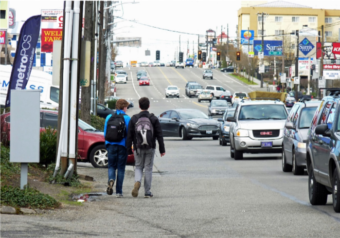 Two kids with backpacks walk along a busy multi-lane roadway