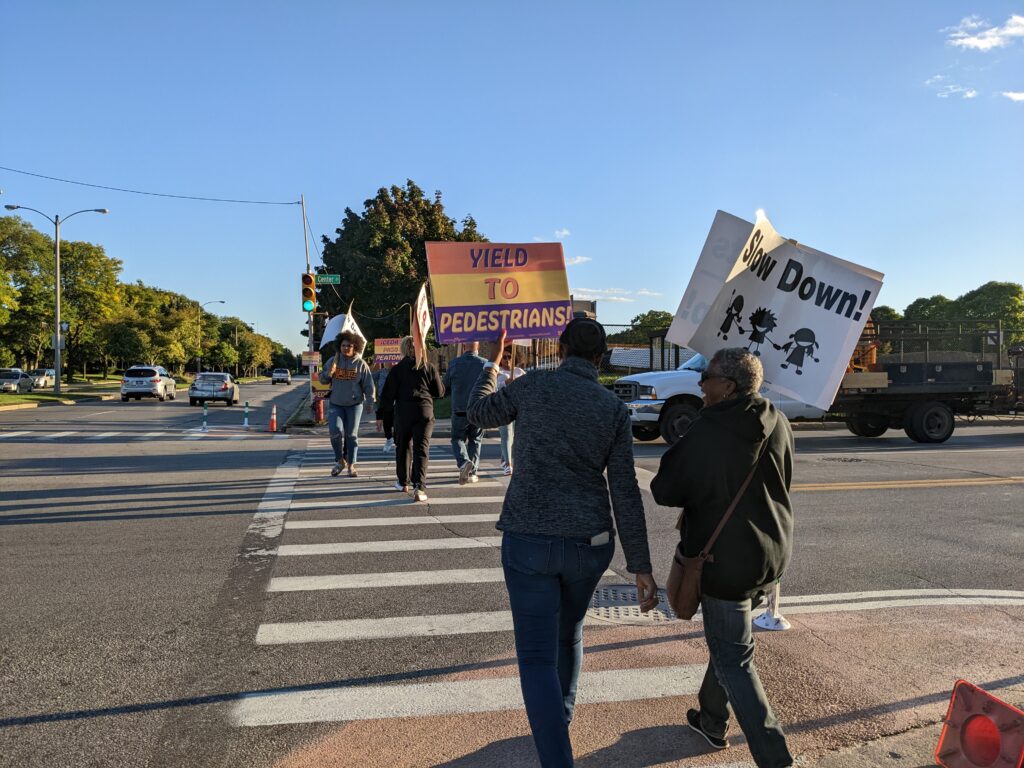 As they cross the street, people hold up signs calling for safer driver behavior, including a brightly-colored sign that says "Yield to pedestrians" and a black-and-white sign showing children holding hands that says "Slow down!"