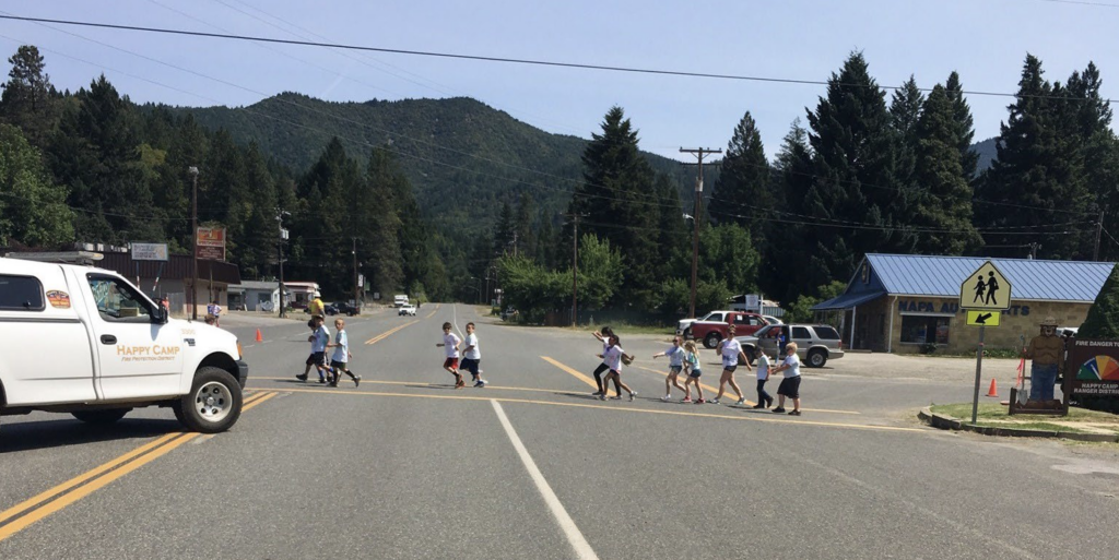 A group of children crosses a large intersection partially blocked by a white pick up truck with the logo "Happy Camp"