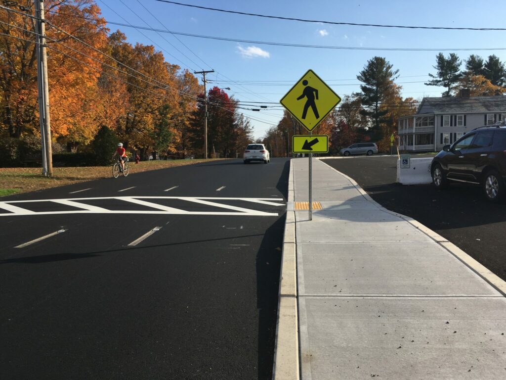 A cyclist travels down fresh, black asphalt, nearing a short crosswalk with a ped crossing sign.