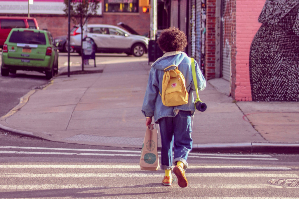 A child with a bright yellow backpack and dark brown, curly hair crosses the street