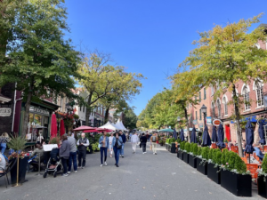 A number of people walks down a shaded street lined with shops and patio seating