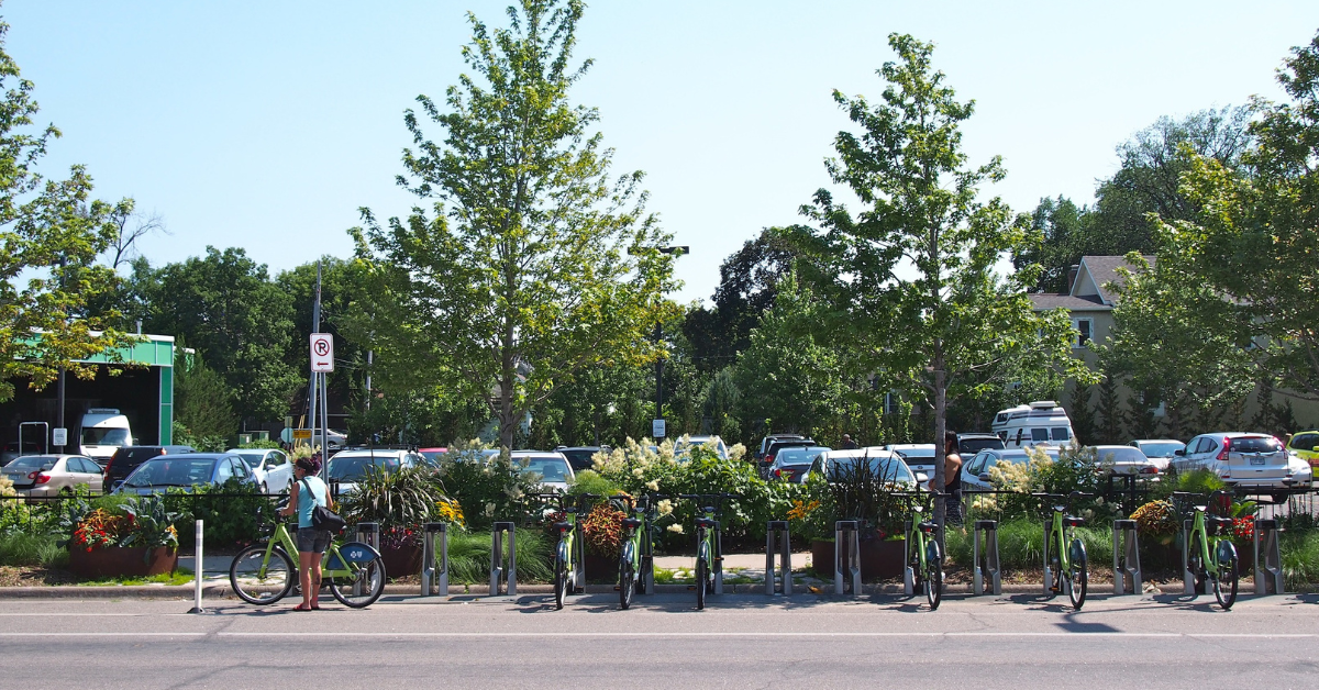 A woman hops on a bicycle beside a row of trees that fails to hide the rows of parking behind her