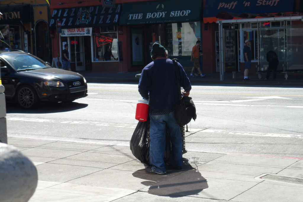 An unhoused person waits at an intersection carrying a bottle of water and two large bags