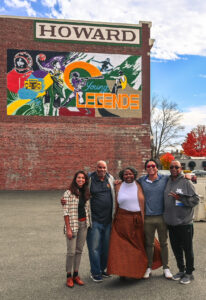 Smart Growth America staff and community leaders pose in front of a colorful mural that features youth playing basketball and engaged in other activities that reads "Young Legends"