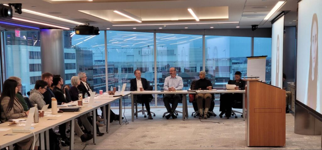 A photo of folks gathered around a table in a cofference room with a woman on teh screen's using the meeting's virtual meeting functions