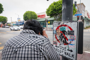 A person looks through the lens of a camera, focusing on a pedestrian crossing sign which has been altered and has the pedestrian image crossed out with a red “no” symbol and other markings.