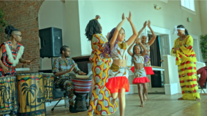 A group of people wearing colorful clothing dance together, some with their arms raised above their heads