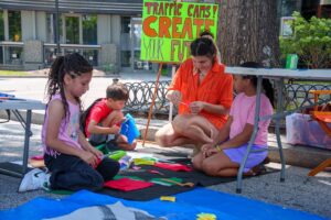 A group of children sit on the ground outdoors, creating a streetscape with a variety of materials.