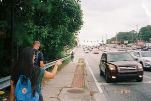 Two people walking on a narrow sidewalk with one person pointing toward the wide, multi-lane road filled with cars.